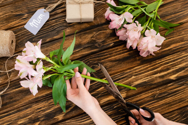 top view of woman holding scissors near pink flowers, gift box and tag with mom lettering, mothers day concept 