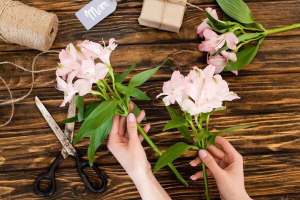 top view of woman holding pink flowers near scissors, jute twine rope and present with mom lettering on tag, mothers day concept 