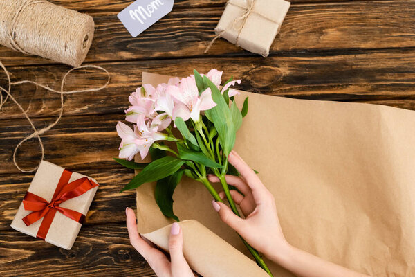 top view of woman holding pink flowers near paper, jute twine rope and gift boxes on wooden surface, mothers day concept 