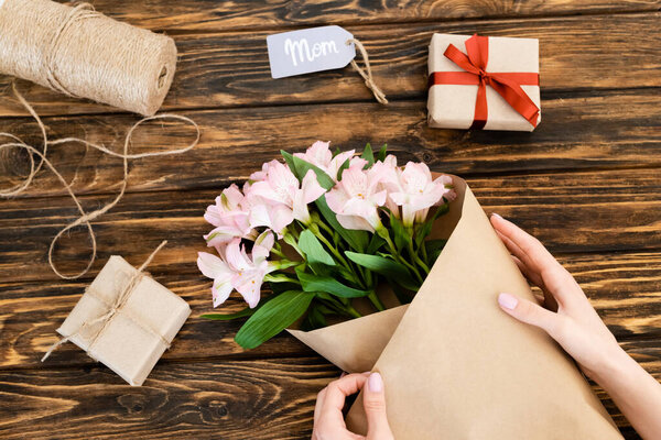 cropped view of woman wrapping pink flowers in paper near gift boxes on wooden surface, mothers day concept 