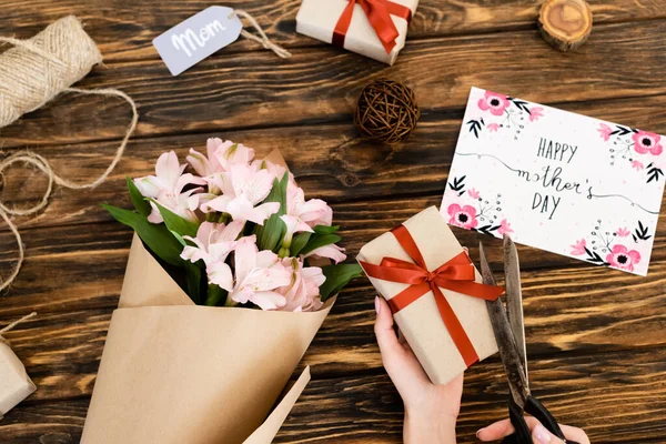 ピンクの花の近くにプレゼントにリボンを切断女性のクロップドビュー 幸せな母の日とグリーティングカードとタグ付きのお母さんのレタリング木製の表面 — ストック写真