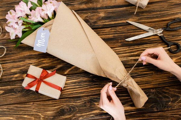 cropped view of woman touching jute twine rope on pink flowers wrapped in paper near gift box and scissors, mothers day concept 