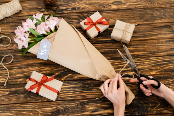 cropped view of woman cutting jute twine rope on pink flowers wrapped in paper near gift boxes and scissors, mothers day concept 