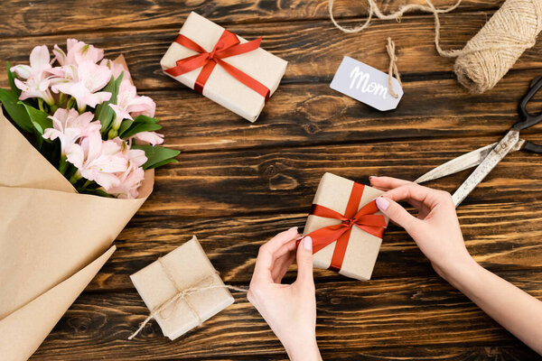 cropped view of woman touching ribbon on present near pink flowers wrapped in paper and tag with mom lettering, mothers day concept 