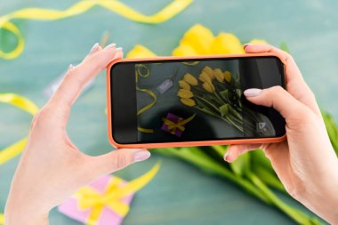 selective focus of woman taking photo of yellow tulips, present and mom tag lettering  on textured surface, mothers day concept  clipart