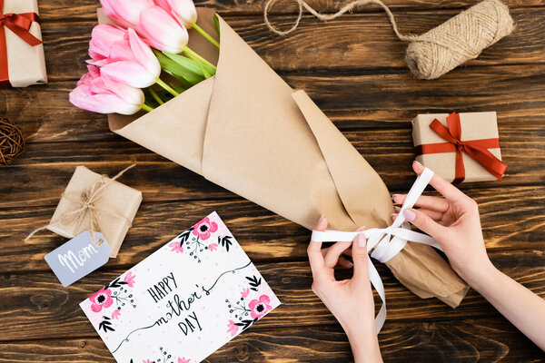 cropped view of woman touching ribbon on wrapped in paper pink tulips near greeting card with happy mothers day lettering and gift boxes on wooden surface  