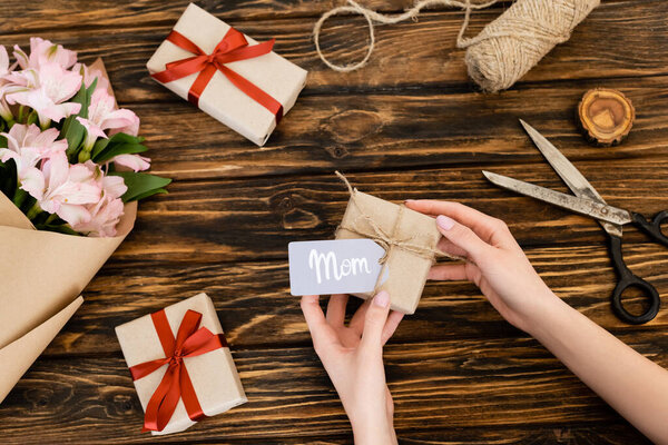 cropped view of woman holding gift box with mom lettering on tag near pink flowers wrapped in paper on wooden surface, mothers day concept 