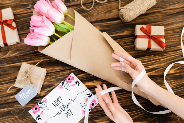 cropped view of woman holding ribbon near wrapped in paper pink tulips, greeting card with happy mothers day lettering and gift boxes on wooden surface  
