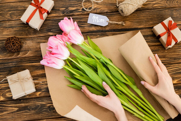 cropped view of woman wrapping pink tulips in paper near gift boxes on wooden surface, mothers day concept 
