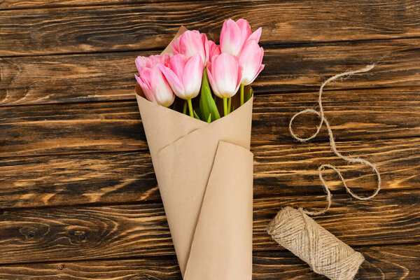 top view of pink tulips wrapped in paper near jute twine rope on wooden surface, mothers day concept 