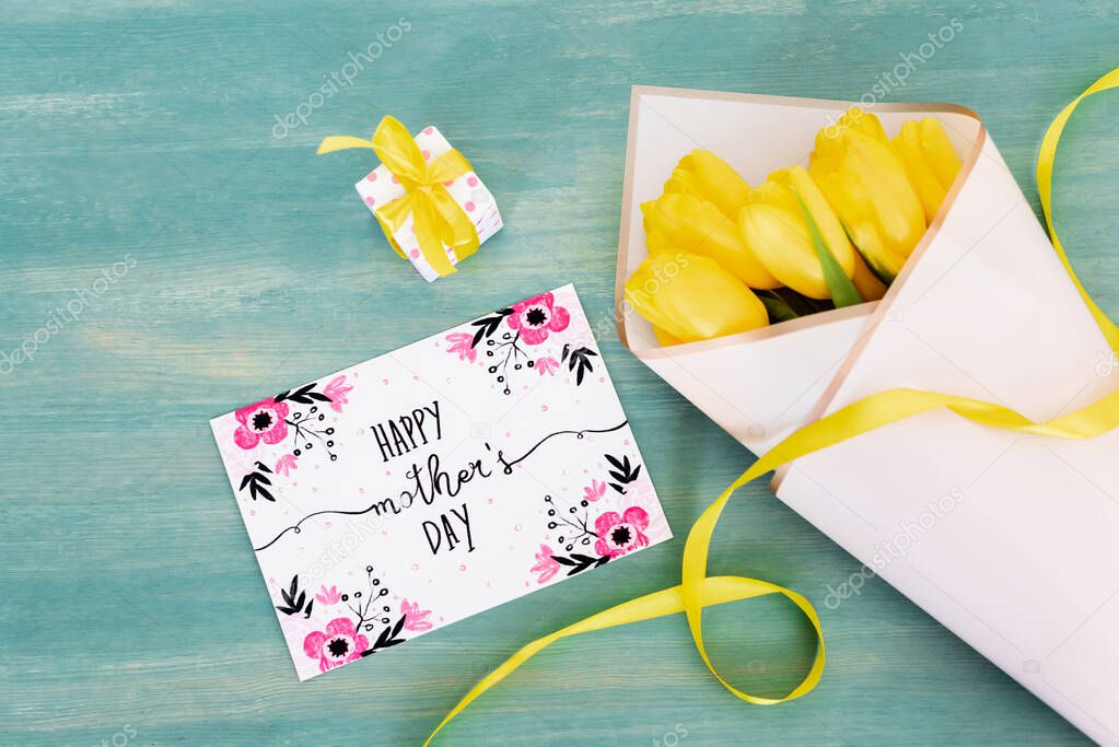top view of small gift box near greeting card with happy mothers day lettering and yellow tulips on blue wooden surface 