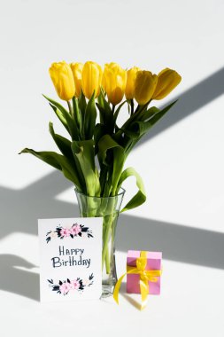 Vazodaki sarı laleler üzerinde güneş ışığı hediye kutusunun yanında ve doğum günü kartı beyaz harflerle.