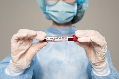 Doktorun elinde kan örneğiyle test tüpü tutuşu ve gri harfli koronavirüs görüntüsü.