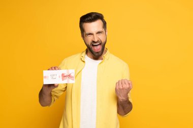 Cheerful man showing yeah gesture and holding gift card on yellow background clipart