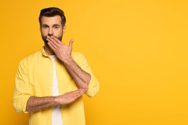 Sarı arka planda işaret dili kullanırken ağzını kapatan yakışıklı bir adam.