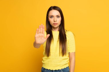Attractive girl showing stop gesture while looking at camera on yellow background clipart