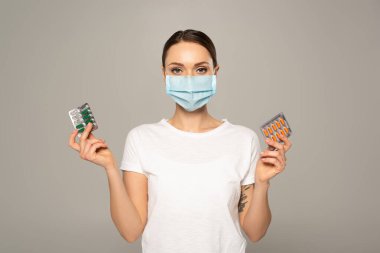 Woman in medical mask holding blisters with pills isolated on grey  clipart