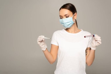 Tıbbi maskeli genç bir kadın, elinde termometre ve test tüpü tutuyordu. Üzerinde kan örneği ve koronavirüs yazıları vardı. 