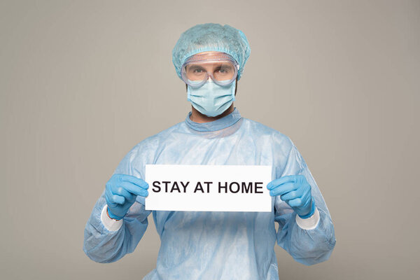 Doctor in medical mask and protective googles holding card with stay at home lettering isolated on grey