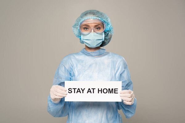 Doctor in medical mask and cap holding card with stay at home lettering isolated on grey