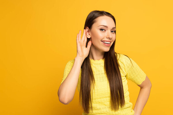 Smiling girl with hand near ear looking at camera on yellow background