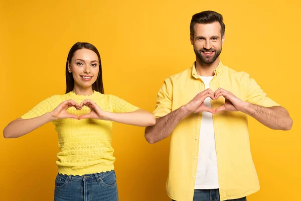 Lächelndes Paar Zeigt Herzenszeichen Und Blickt Die Kamera Auf Gelbem — Stockfoto