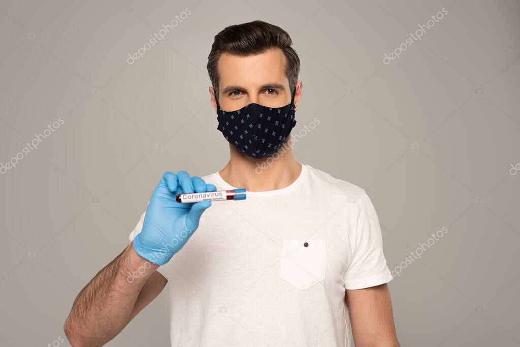 Man in safety mask and latex glove holding test tube with blood sample and coronavirus lettering isolated on grey 