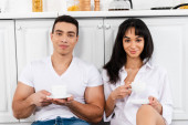 Mezirasový pár s podšálky a šálky kávy s úsměvem a při pohledu do kamery v blízkosti kuchyňských skříněk