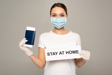 Woman in medical mask holding passport with air ticket and card with stay at home lettering isolated on grey  clipart