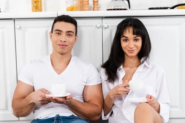 Interracial Couple Saucers Cups Coffee Smiling Looking Camera Kitchen Cabinets — Stock Photo, Image