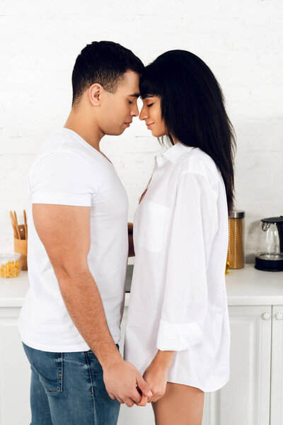 Interracial couple with closed eyes facing each other and holding hands in kitchen