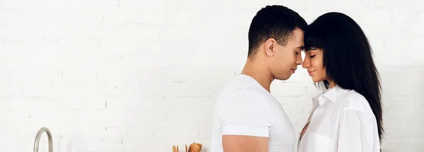 Interracial couple with closed eyes facing each other and smiling in kitchen, panoramic shot