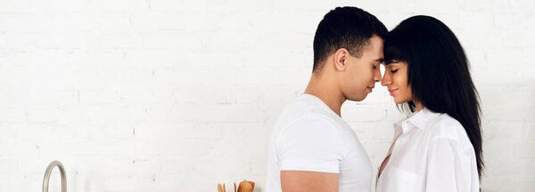 Interracial couple with closed eyes facing each other and smiling in kitchen, panoramic shot