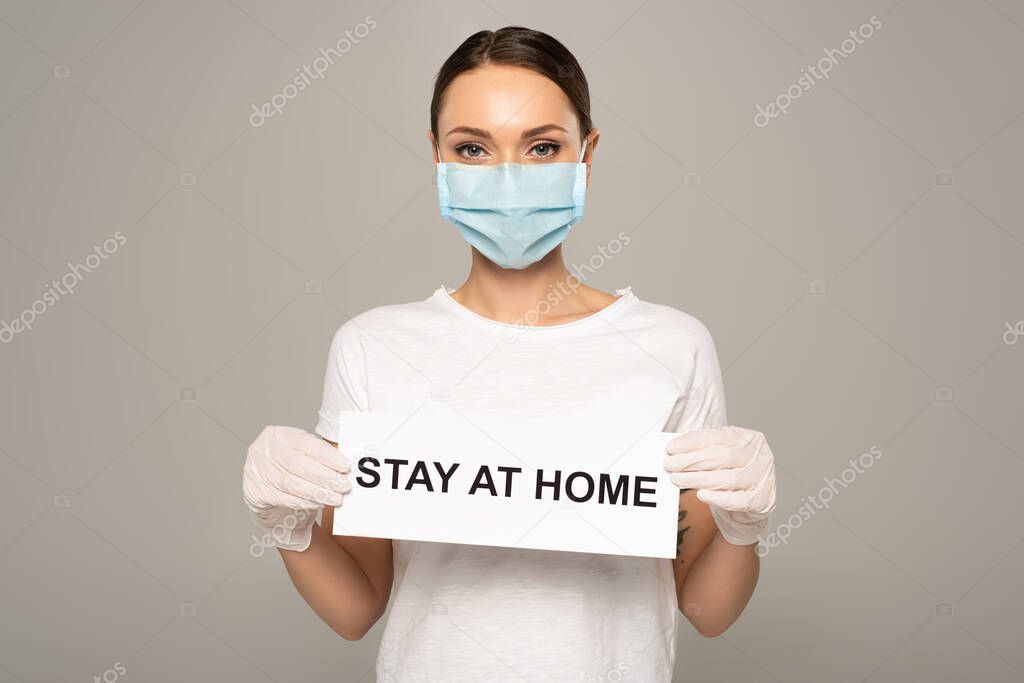 Young woman in medical mask and latex gloves holding card with stay at home lettering isolated on grey 