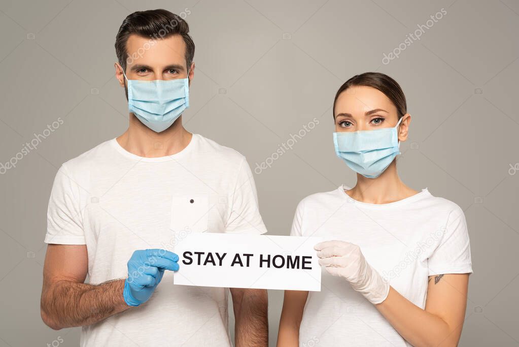 Young couple in medical masks holding card with stay at home lettering isolated on grey 