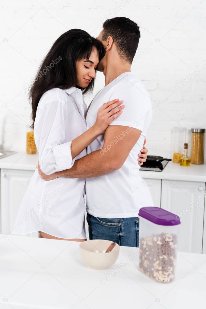 African american girl with closed eyes smiling and hugging with boyfriend in kitchen
