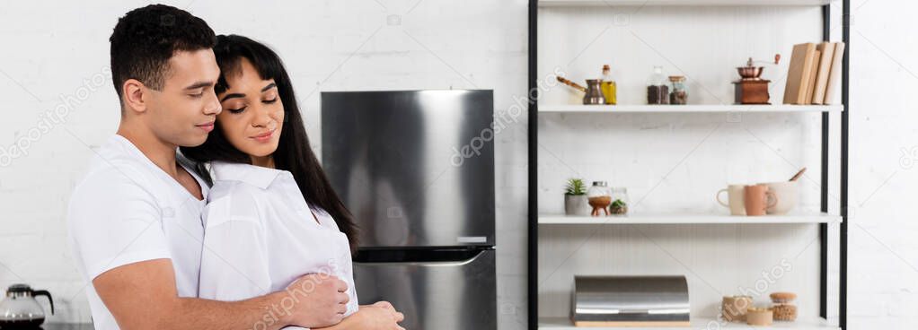 Side view of man hugging african american girl with closed eyes from behind in kitchen, panoramic shot