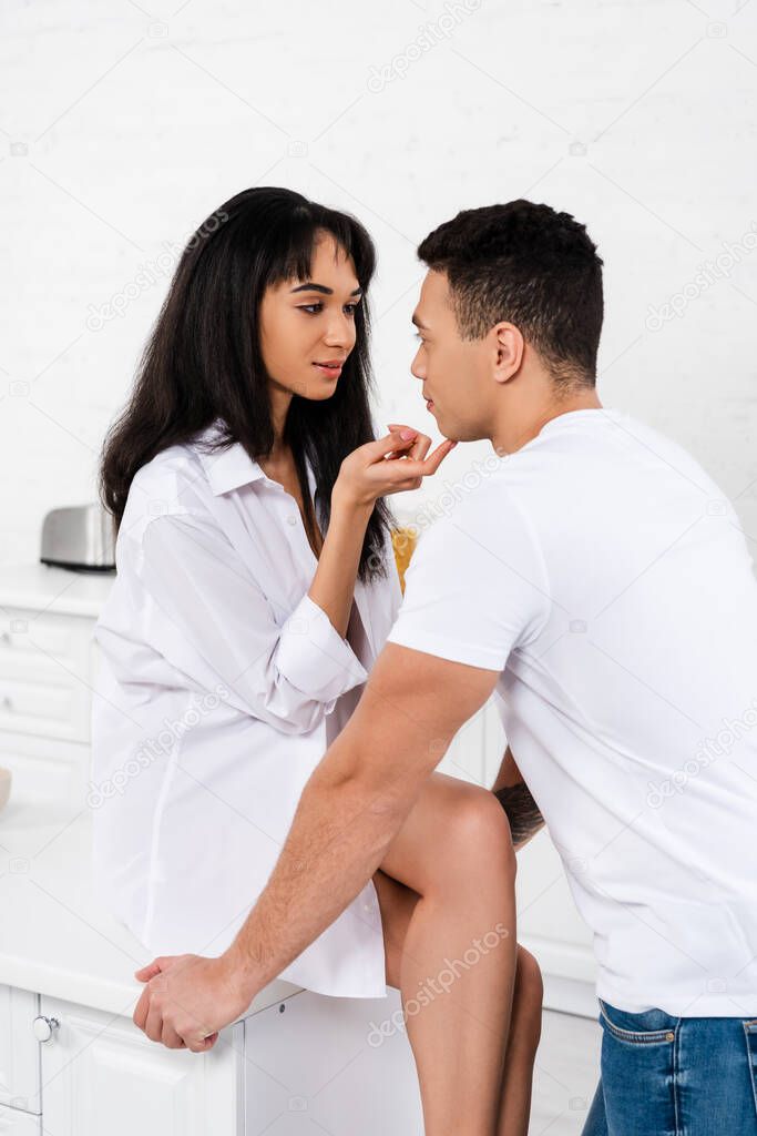 African american woman sitting on table, smiling and looking at boyfriend in kitchen