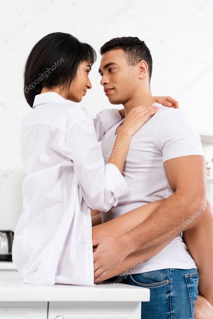 Interracial couple looking at each other and african american woman sitting on table and hugging with boyfriend in kitchen