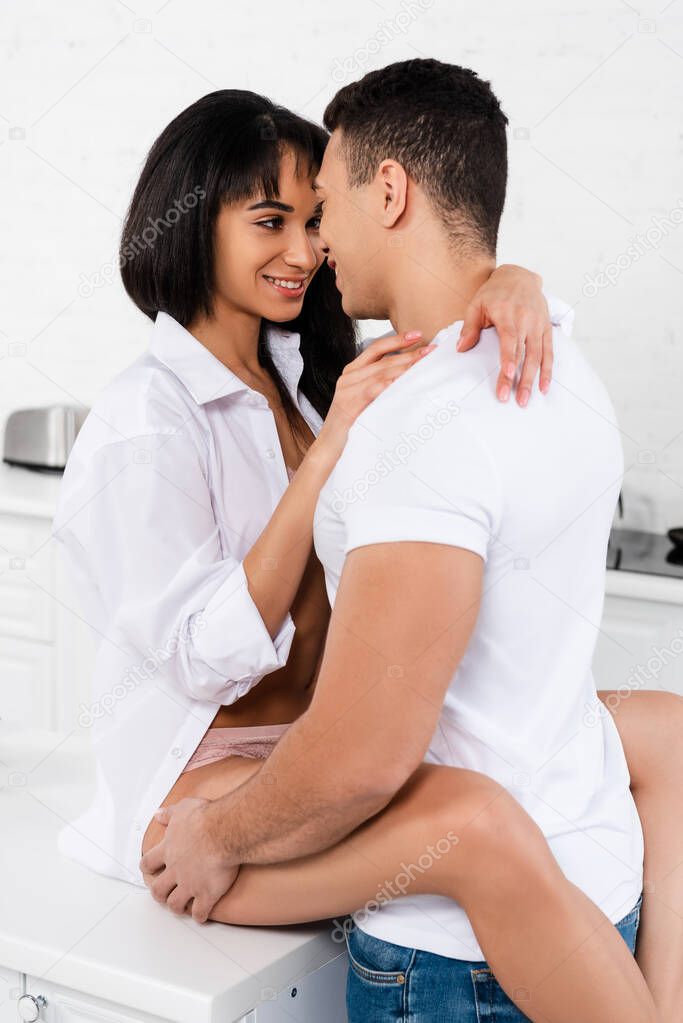 Interracial couple looking at each other, smiling and african american woman on table hugging with boyfriend in kitchen