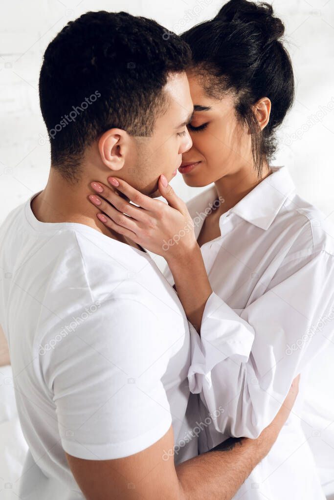 Interracial couple hugging and kissing with closed eyes in bedroom