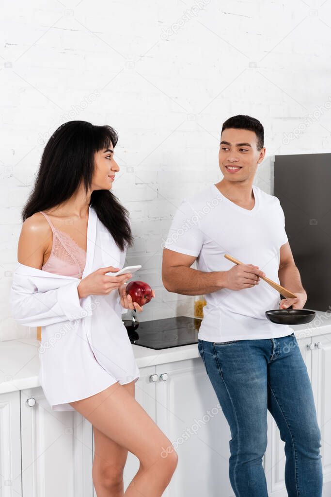 Interracial couple with frying pan, spatula, smartphone and apple looking at each other and smiling in kitchen