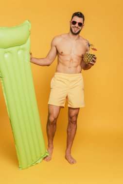 Şişme şilte tutan neşeli kaslı adam ve sarı arka planda taze kokteyl olan yarım ananas.