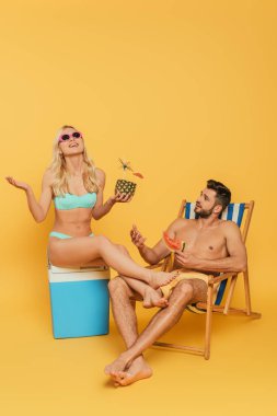 excited blonde girl sitting on portable fridge near handsome man holding slice of watermelon in deckchair on yellow background clipart