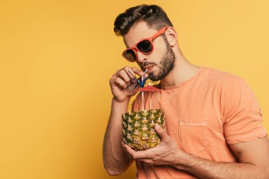 confident young man in sunglasses drinking fresh cocktail from half of fresh pineapple on yellow background clipart
