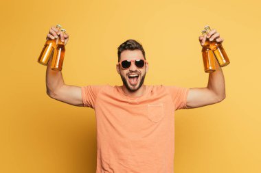 Sarı arka plandaki kameraya bakarken elinde bira şişeleri olan heyecanlı genç adam.