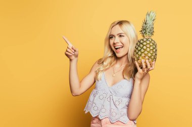 Ananas tutan ve sarı arka planı işaret eden neşeli sarışın kız.