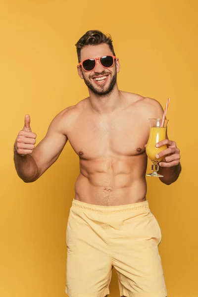 兴奋而强壮的男人戴着太阳镜 拿着装有鲜橙汁的鸡尾酒杯 背对着黄色的背景 露出大拇指 — 图库照片