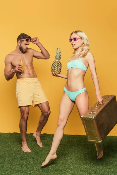 ショック男タッチ眼鏡で魅力的なブロンドの女の子を見ながら水着で歩くとともにヴィンテージスーツケースの上に黄色の背景 — ストック写真