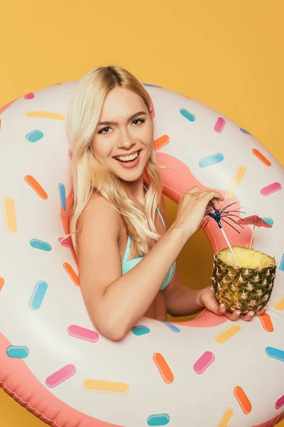 happy blonde girl in swim ring holding half of pineapple with fresh cocktail on yellow background